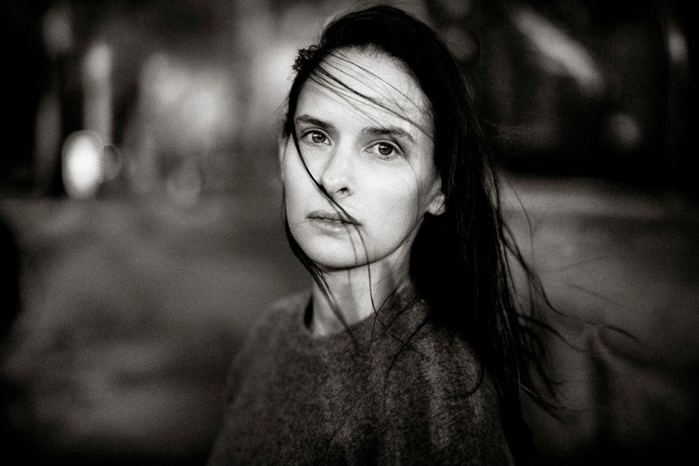Black and white portrait of a young woman with dark hair blowing across her face outdoors.