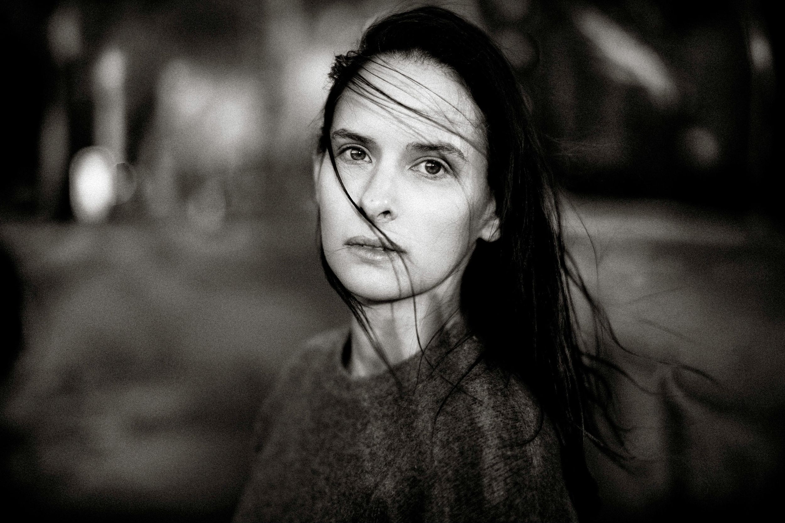 Black and white portrait of a young woman with dark hair blowing across her face outdoors.