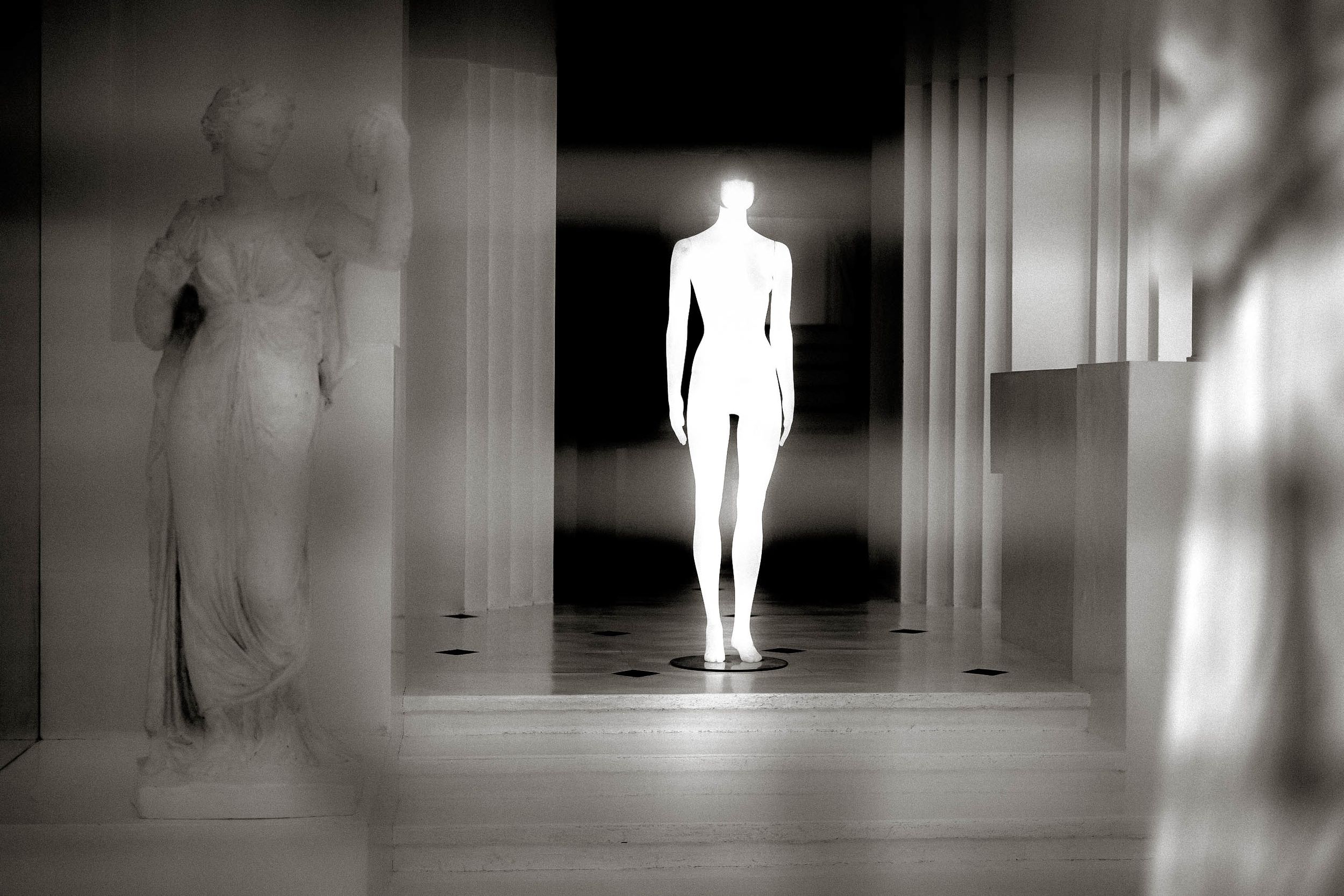 Glowing white humanoid figure stands between classical sculptures in a dramatic black and white artistic installation.