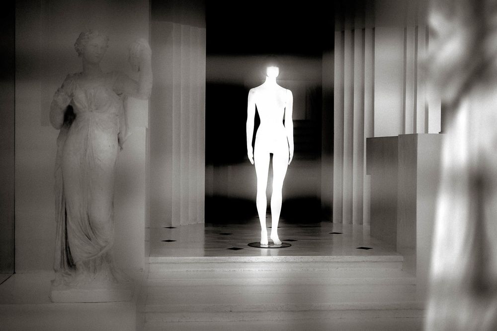 Glowing white humanoid figure stands between classical sculptures in a dramatic black and white artistic installation.