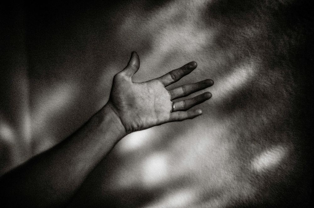 Black and white photo of an outstretched hand reaching forward with light patterns on the wall behind it.