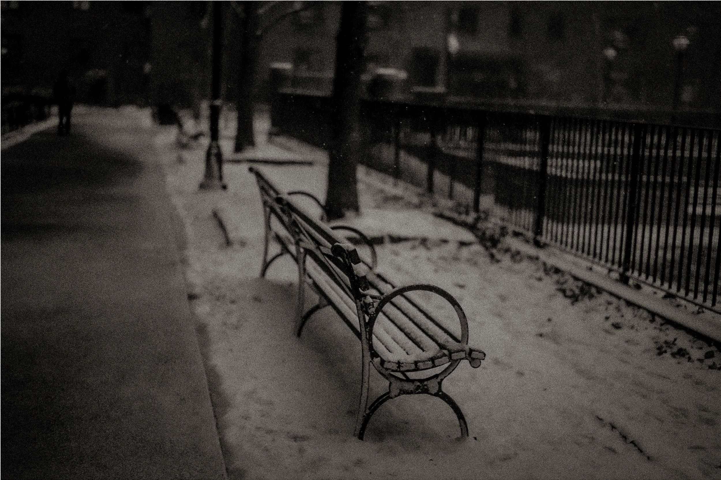 Snow-covered iron park bench along a tree-lined sidewalk with black metal fence in winter.