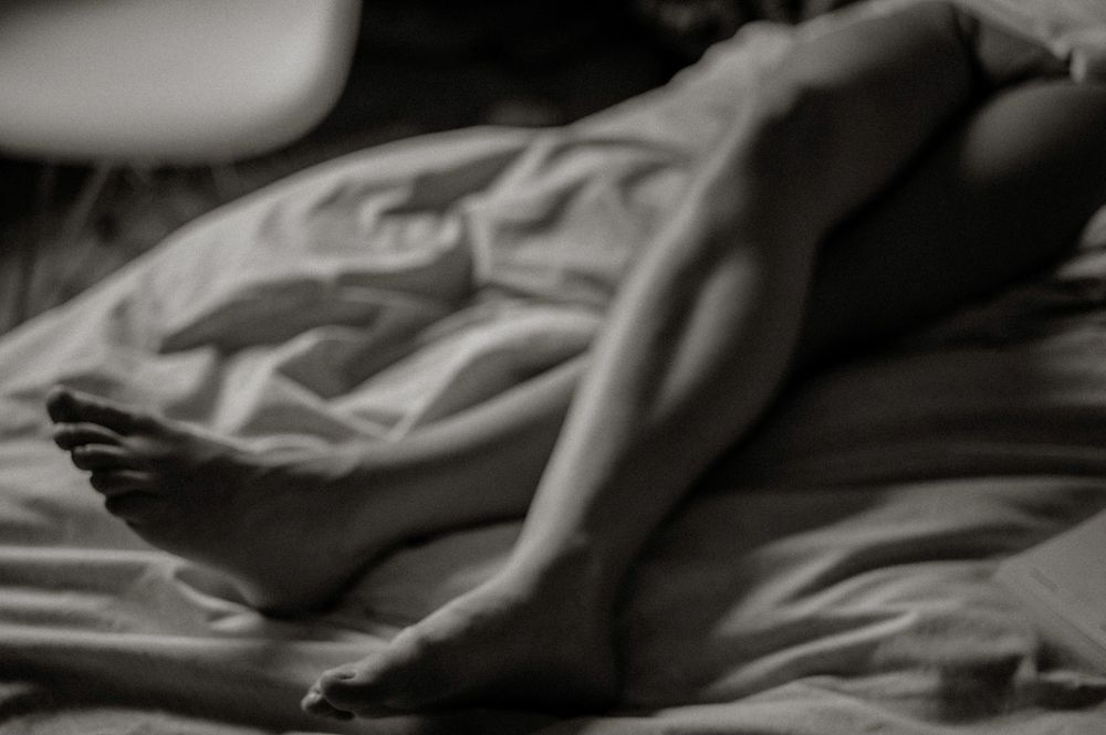 Black and white artistic photo of bare legs resting on rumpled white bed sheets in soft natural light.