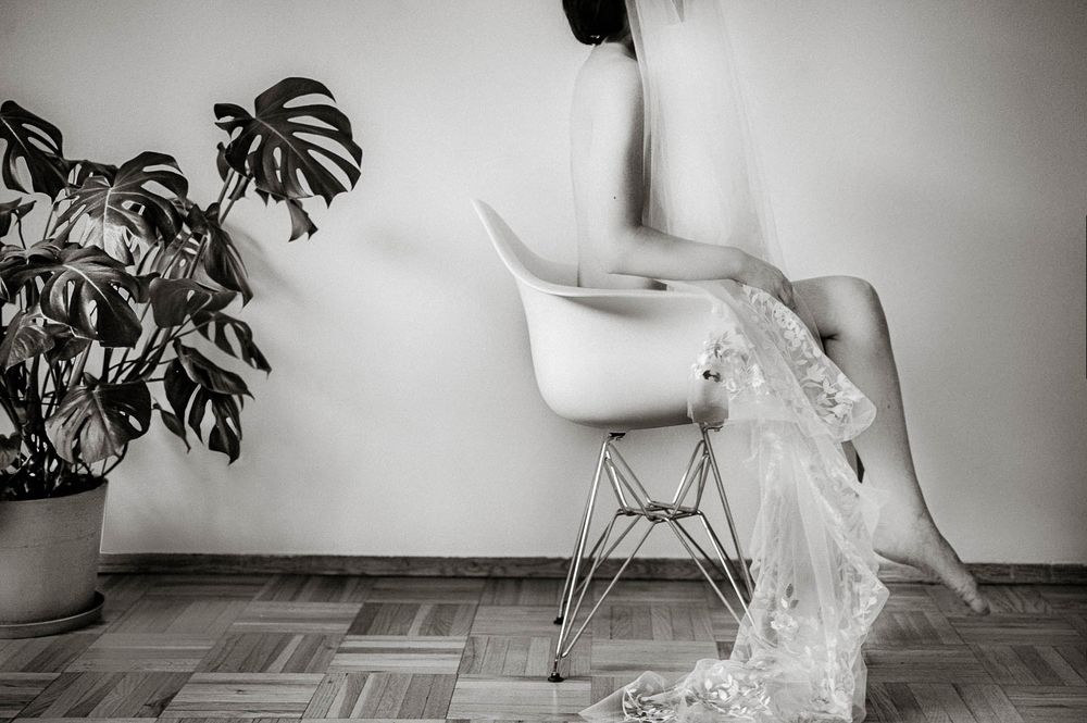 Black and white photo of a person sitting on a modern white chair beside a monstera plant on a wooden floor.