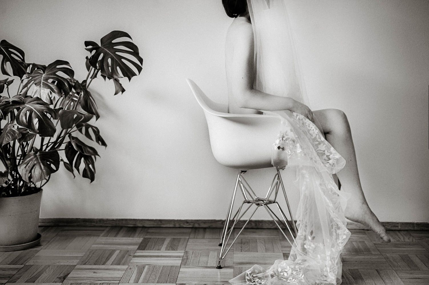 Black and white photo of a person sitting on a modern white chair beside a monstera plant on a wooden floor.
