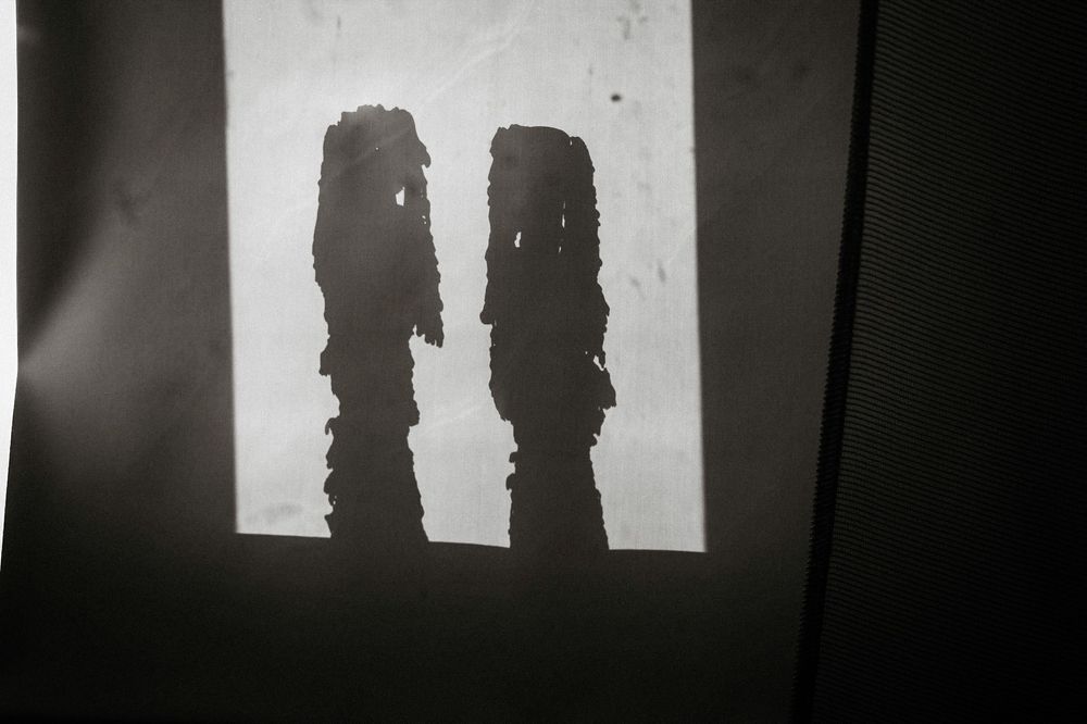 Two dark silhouettes resembling tall figures cast against a bright white wall in a black and white photograph.