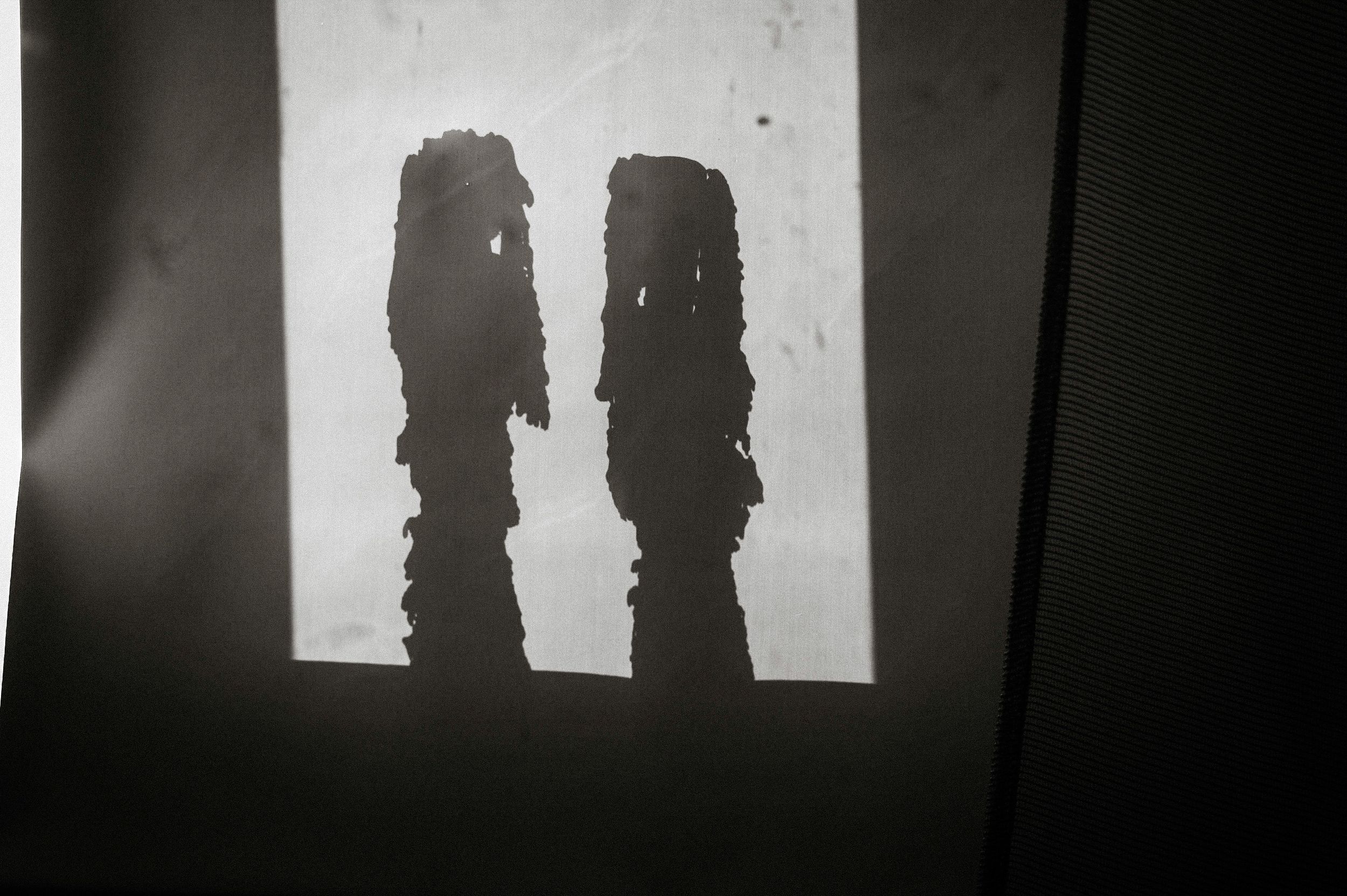 Two dark silhouettes resembling tall figures cast against a bright white wall in a black and white photograph.