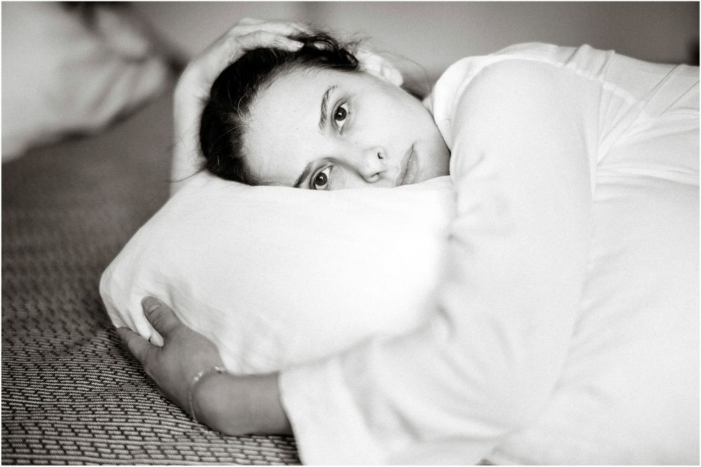 Black and white photo of a young woman lying on a bed, hugging a white pillow and gazing at the camera.
