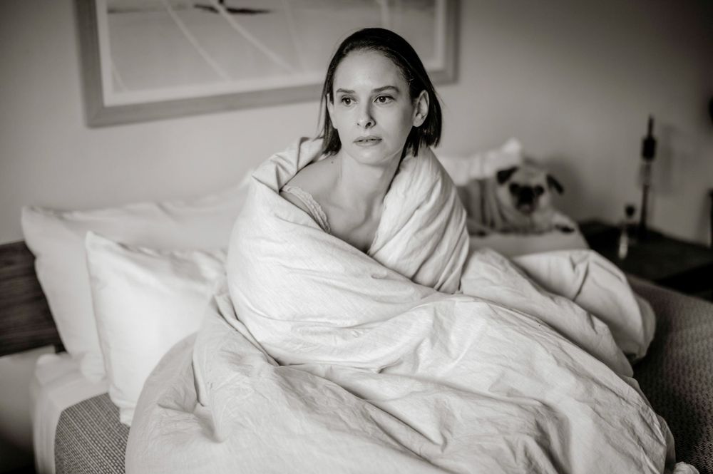 Woman wrapped in white duvet on bed in black and white photo with pug dog in background.