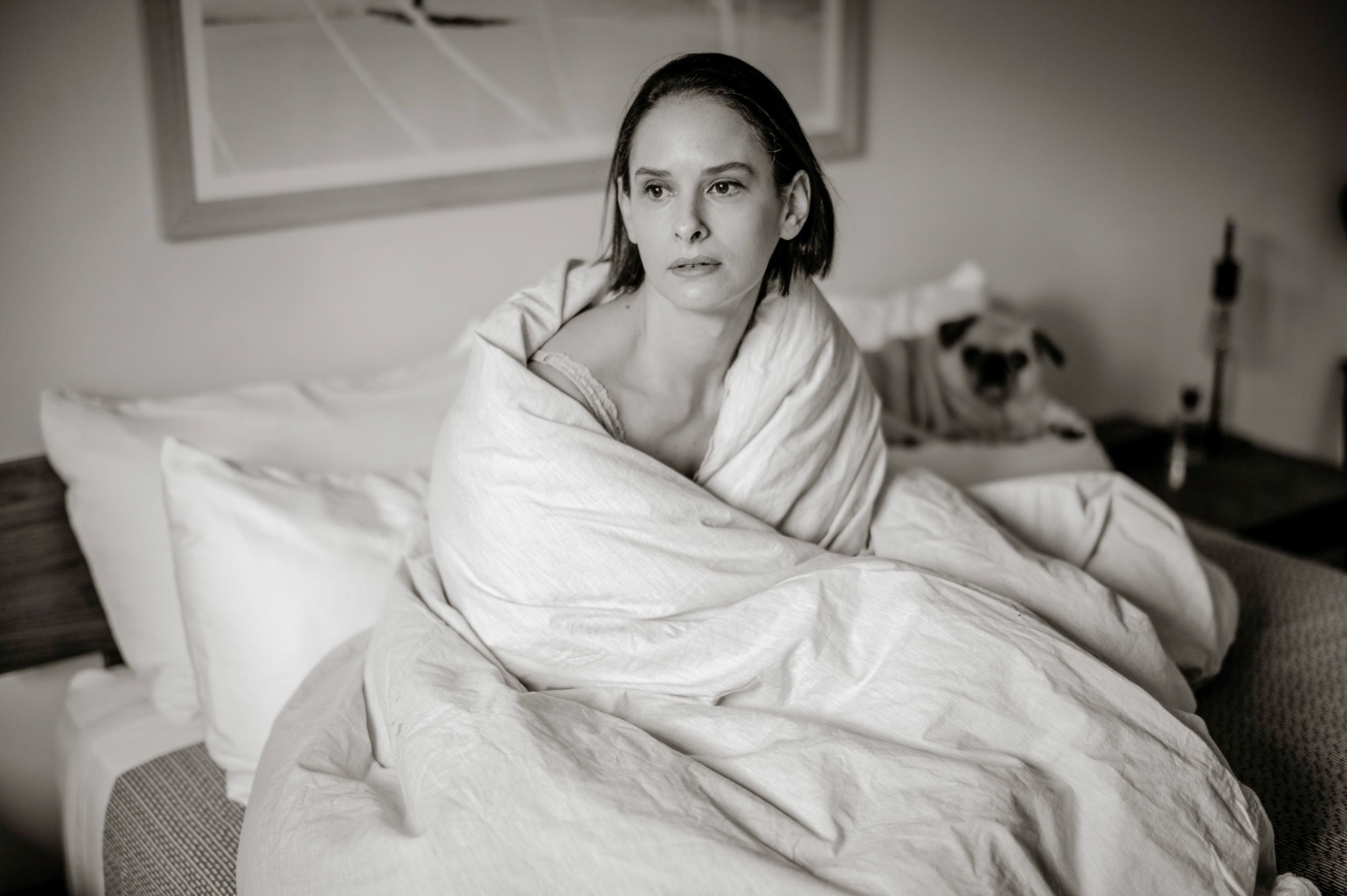 Woman wrapped in white duvet on bed in black and white photo with pug dog in background.