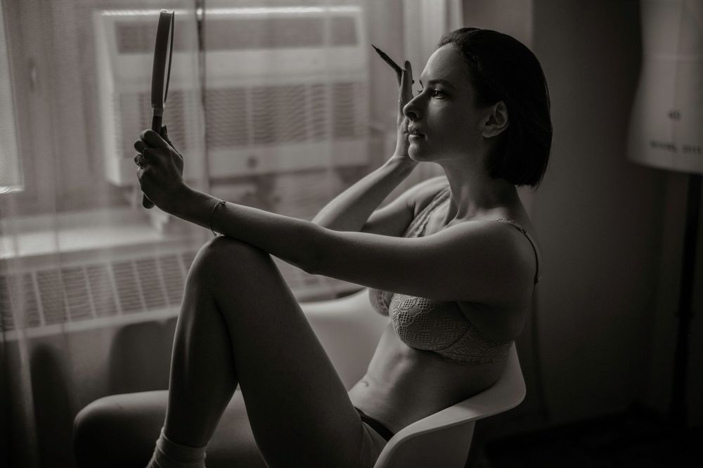 Black and white photo of a woman in lingerie sitting by a window holding a mirror in soft natural light.
