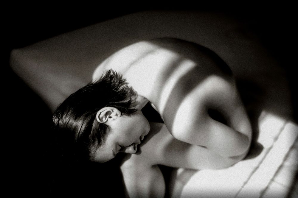 Black and white artistic nude photograph with dramatic light and shadow stripes across a reclining figure.