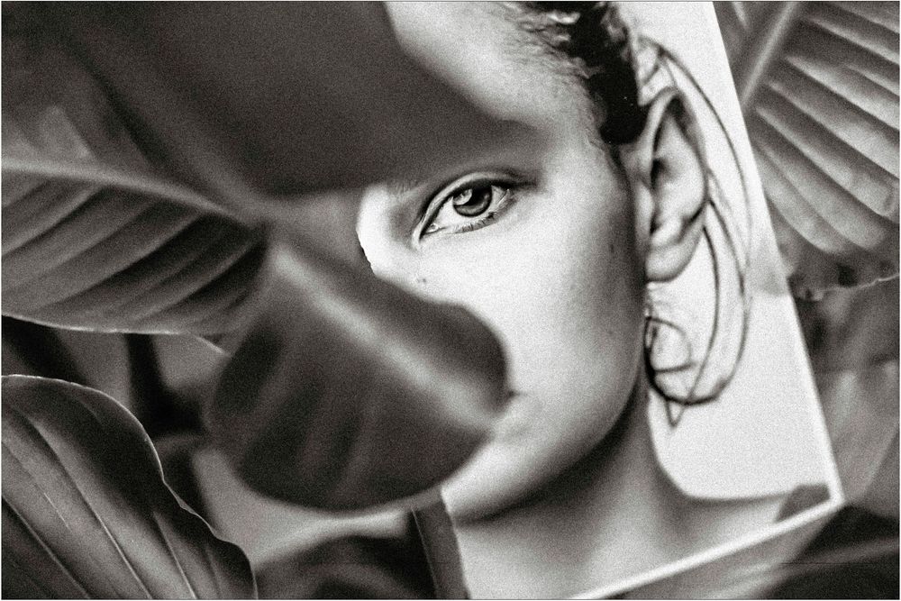 Black and white portrait of a woman partially hidden behind tropical leaves, one eye visible.