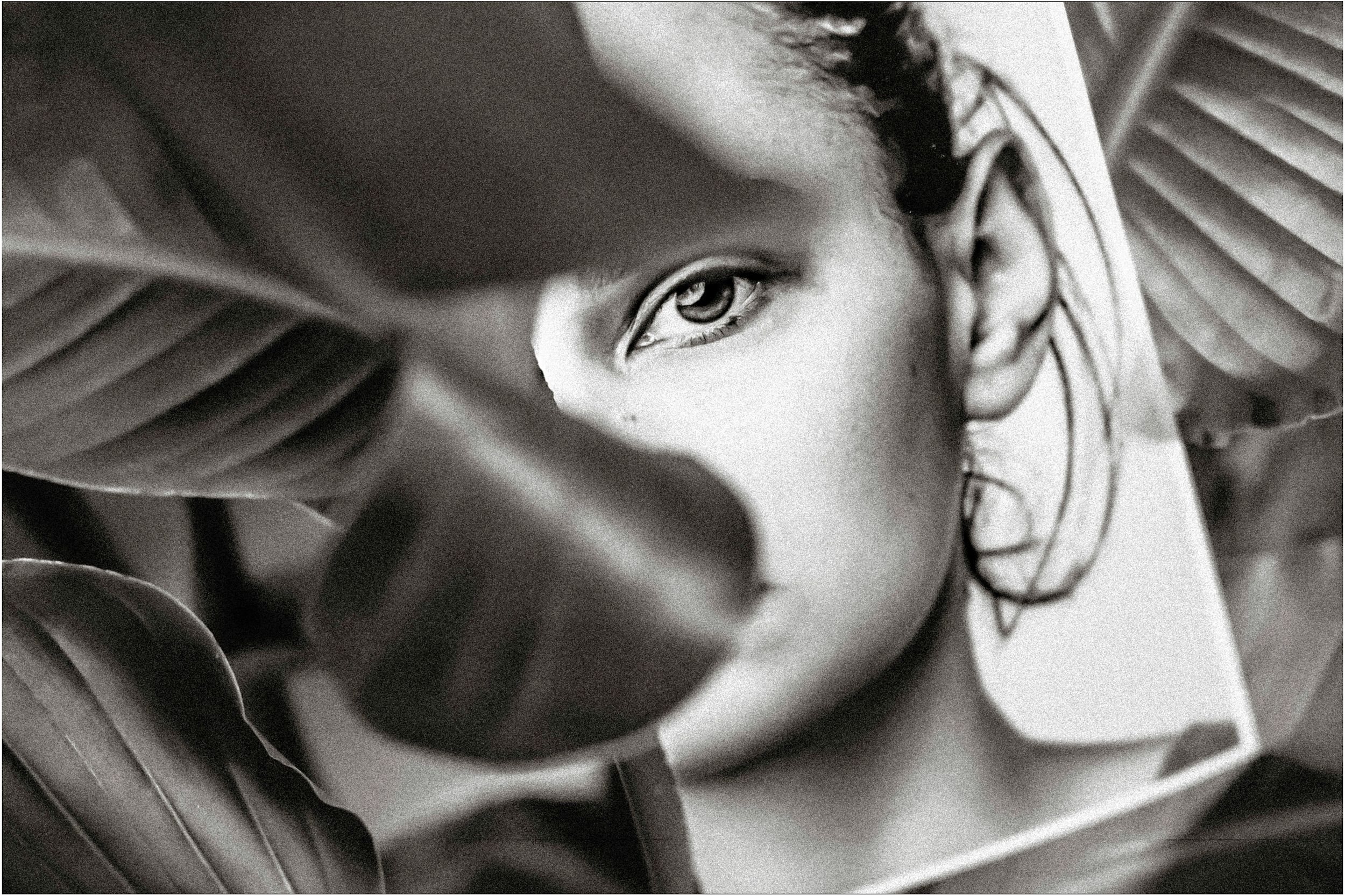 Black and white portrait of a woman partially hidden behind tropical leaves, one eye visible.