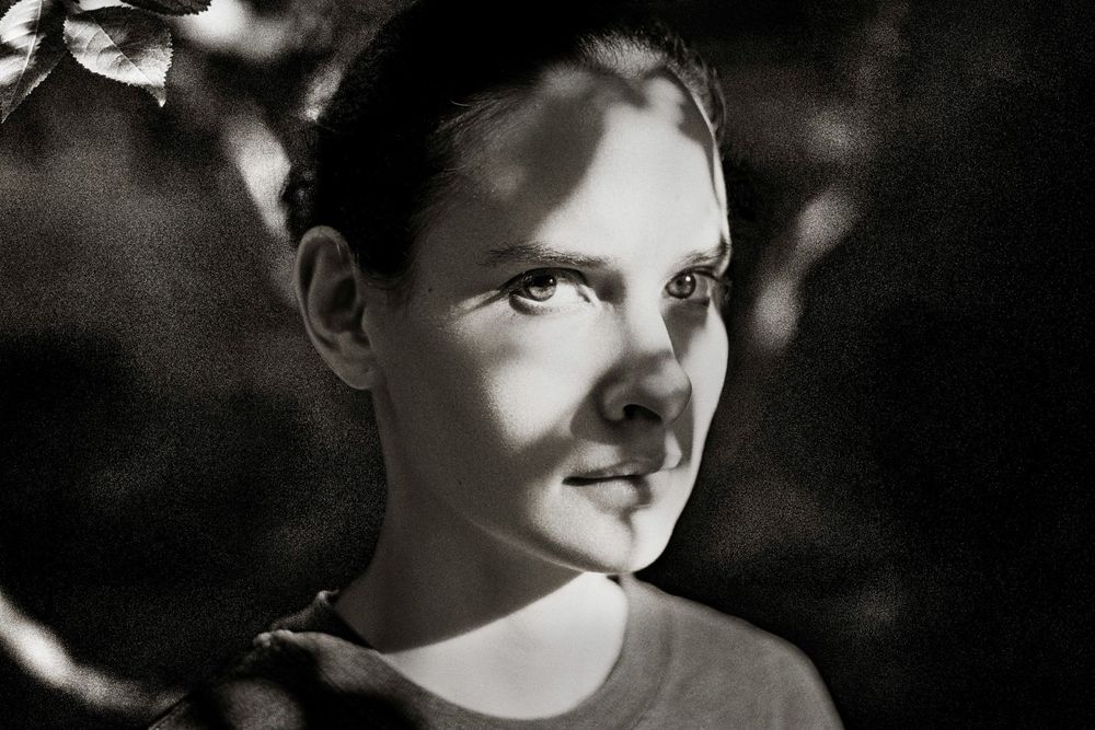 Black and white portrait of a young woman with dappled light and shadow, gazing upward outdoors.