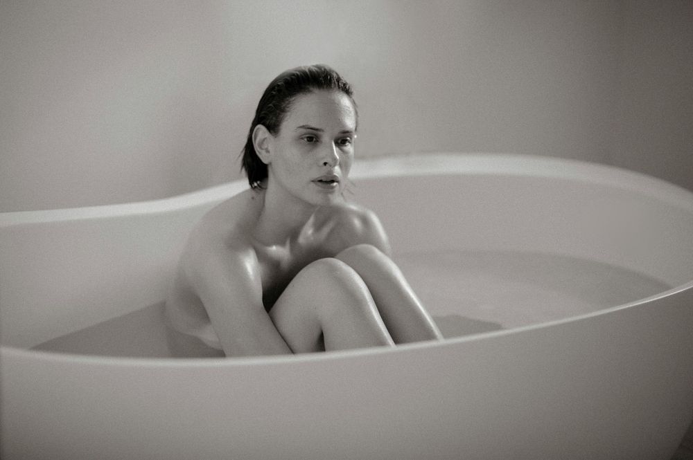 Black and white photo of a woman sitting in a modern freestanding bathtub with knees drawn up.