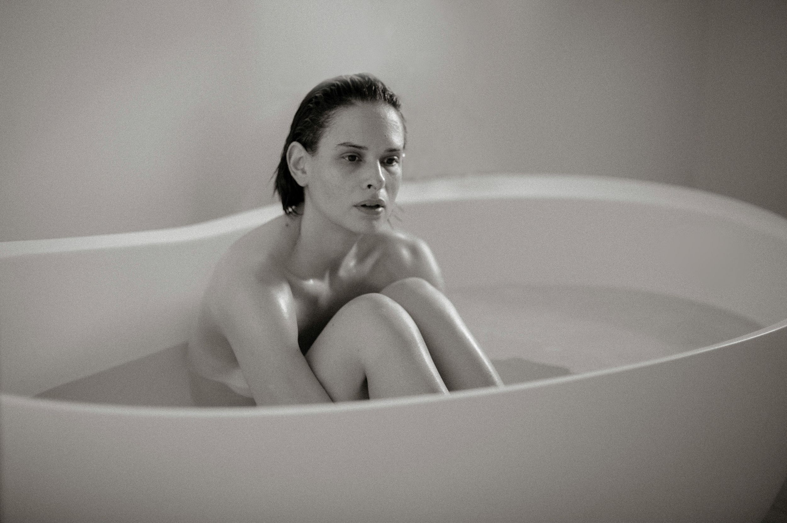 Black and white photo of a woman sitting in a modern freestanding bathtub with knees drawn up.