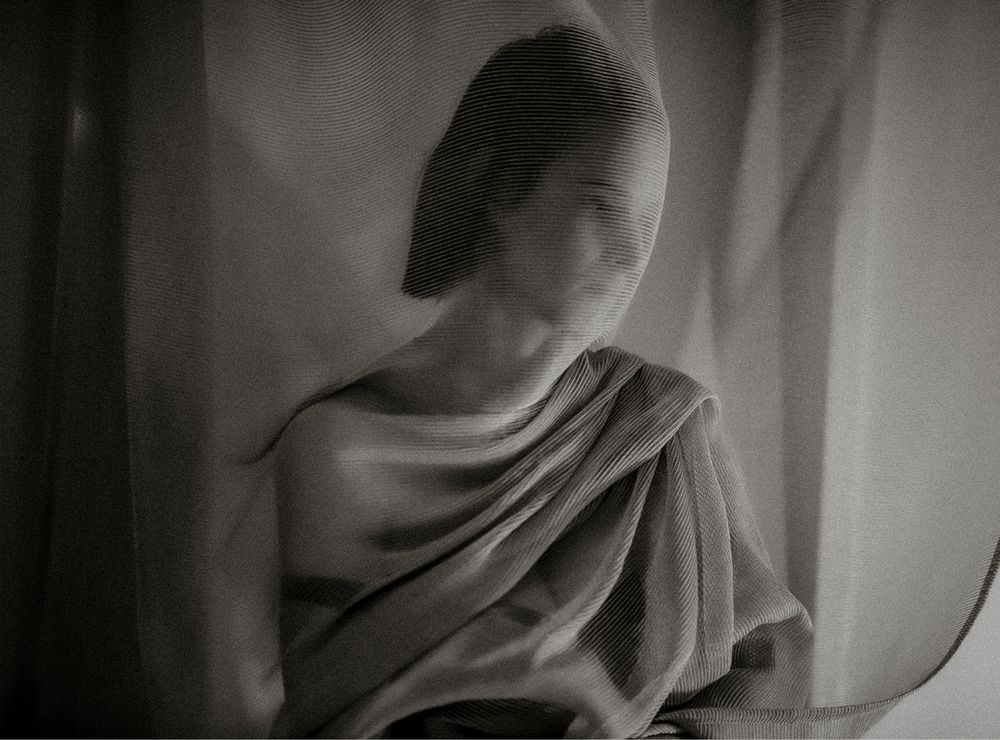 Black and white portrait of a woman draped in fabric, gazing pensively by a window in soft light.