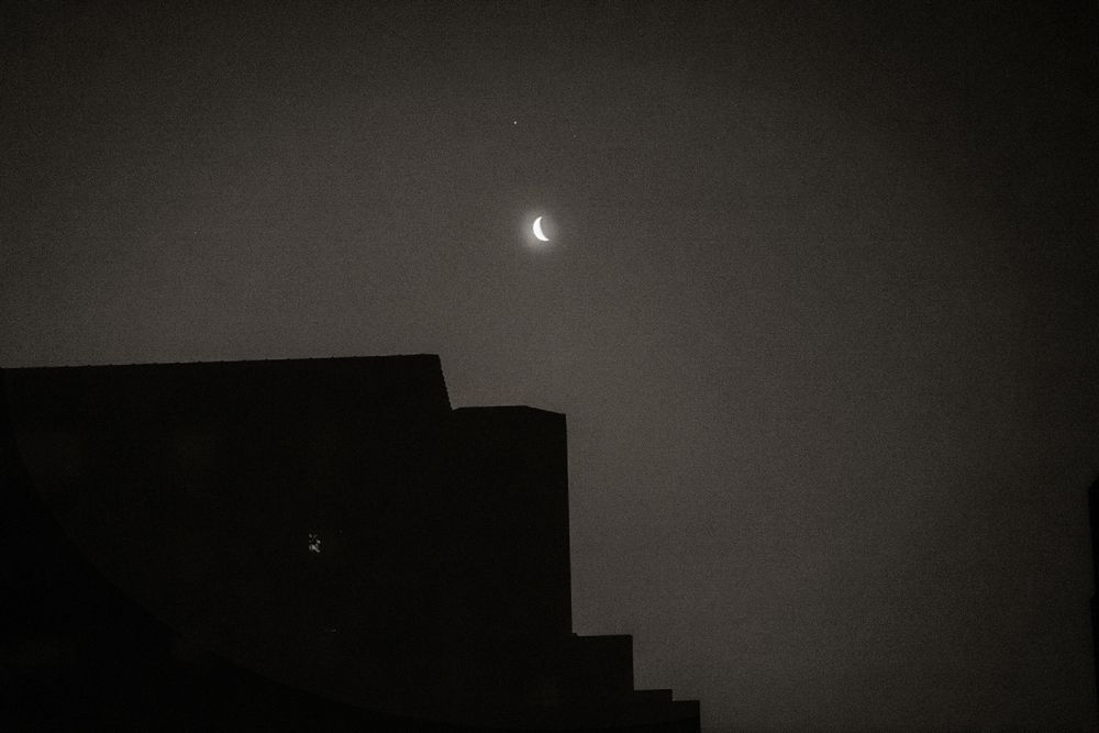 Crescent moon glowing in dark night sky above silhouetted building rooftop.