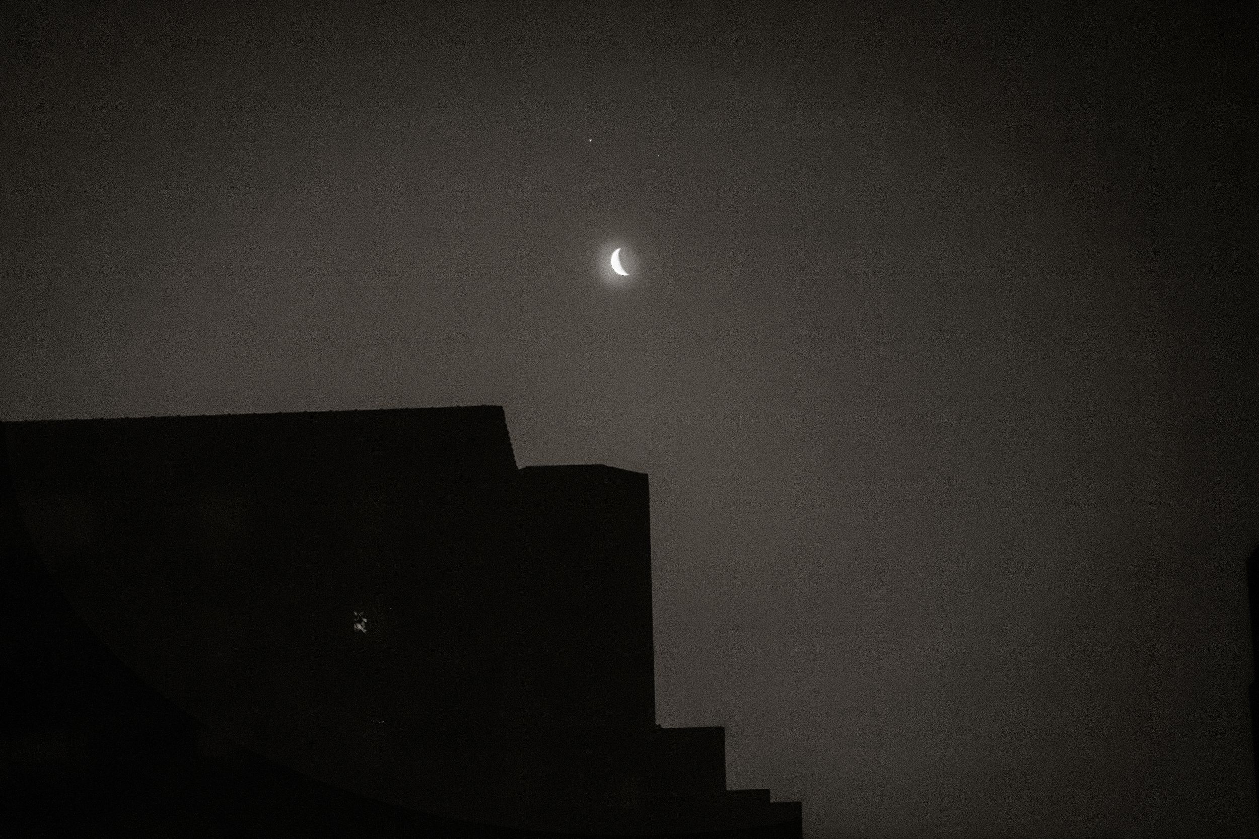Crescent moon glowing in dark night sky above silhouetted building rooftop.