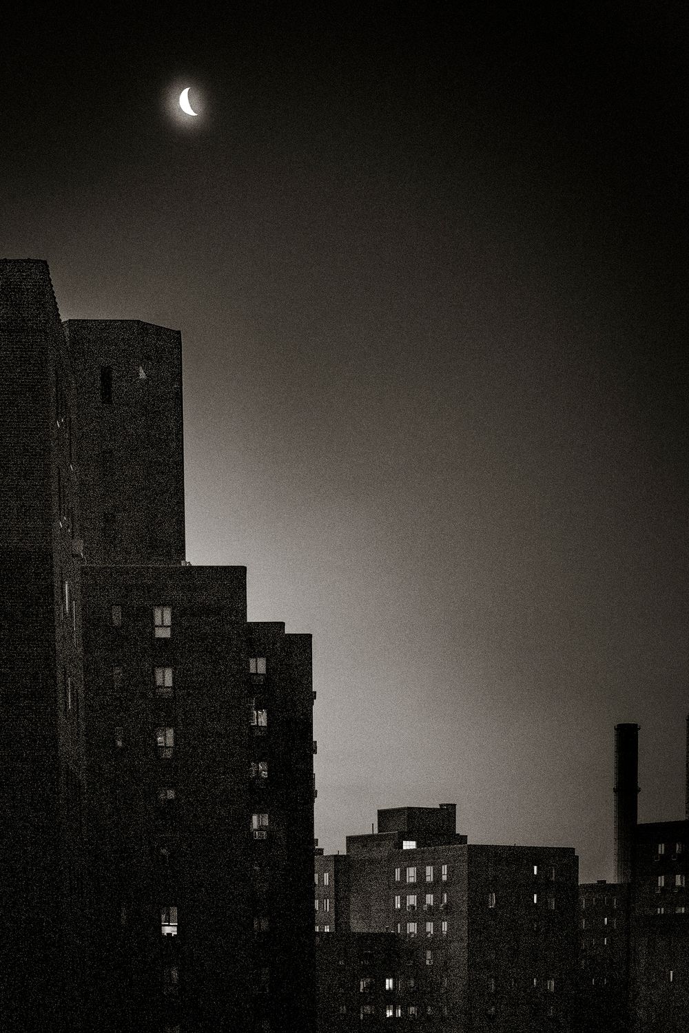 Crescent moon glowing over dark urban skyline at night with silhouetted apartment buildings in black and white.