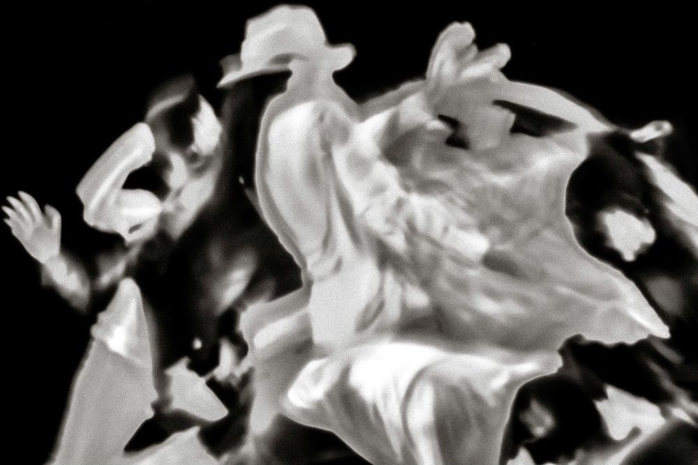 Black and white blurred motion photograph of dancers performing energetically in flowing white costumes.