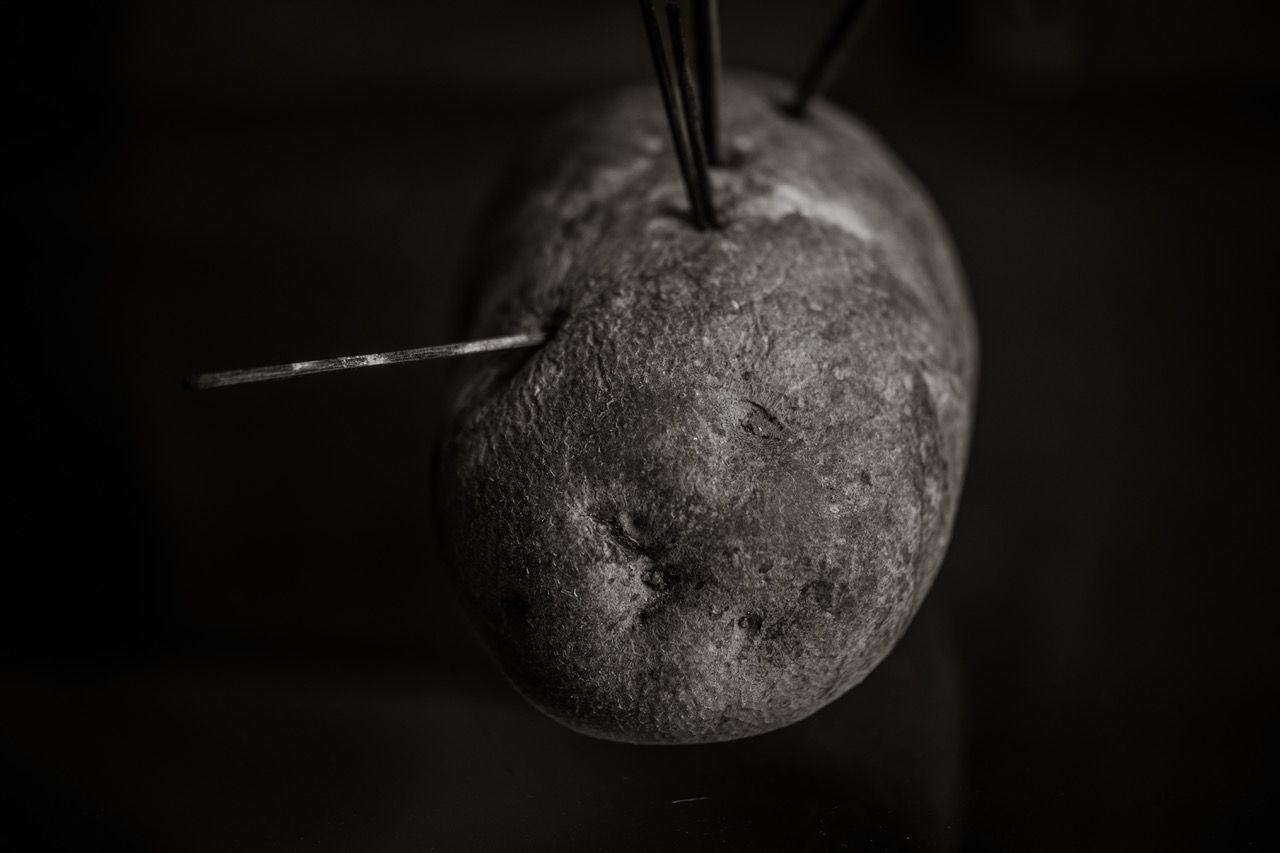 Close-up of a dark, textured potato with metal skewers inserted, captured in dramatic black and white lighting.