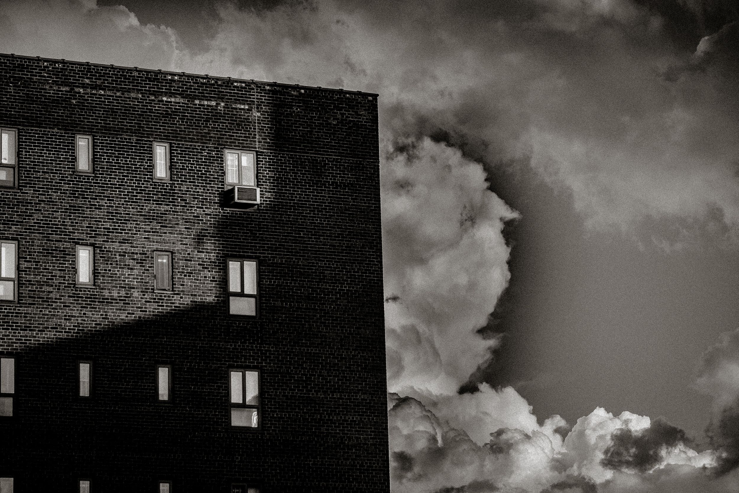 Black and white photo of a brick building beside dramatic storm clouds in the sky.