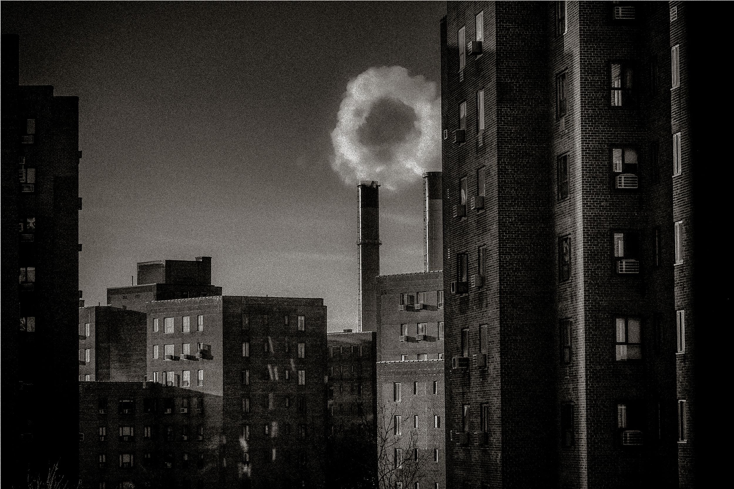 Black and white urban skyline with a smoke stack emitting a circular cloud between dark apartment buildings at dusk.