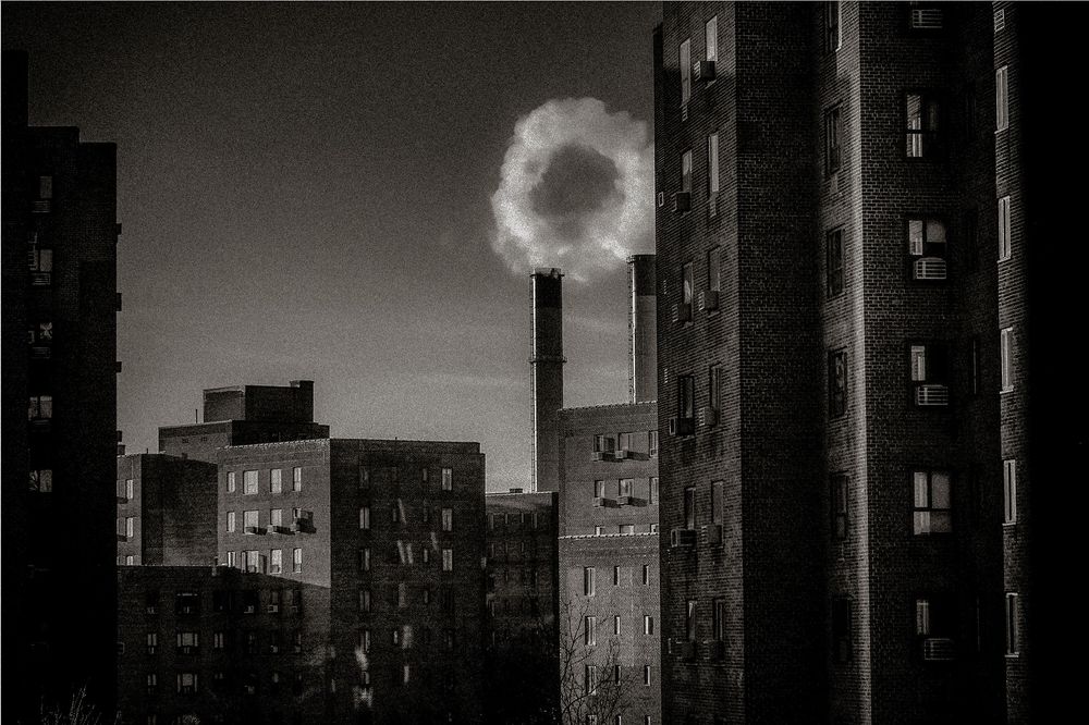 Black and white urban skyline with a smoke stack emitting a circular cloud between dark apartment buildings at dusk.