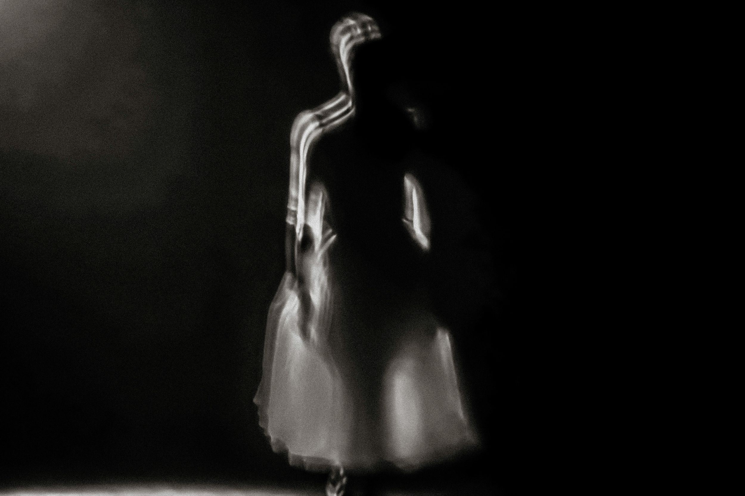 Ghostly blurred figure in white dress standing in dramatic dark spotlight, ethereal long exposure photography.