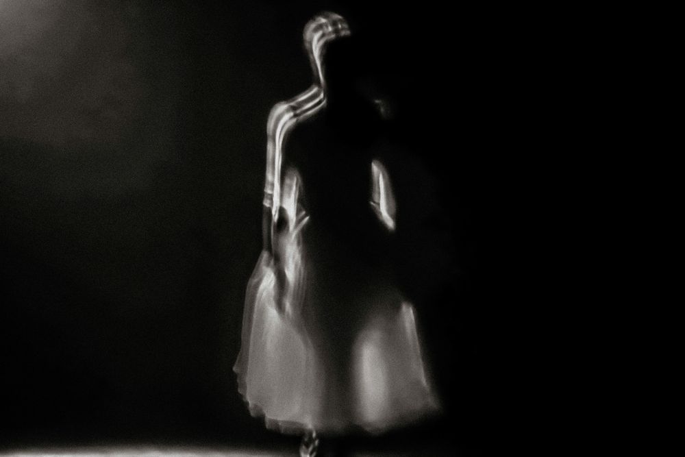 Ghostly blurred figure in white dress standing in dramatic dark spotlight, ethereal long exposure photography.