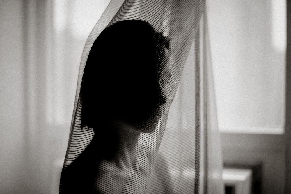 Silhouette of a person with short hair behind sheer curtain in black and white photography.