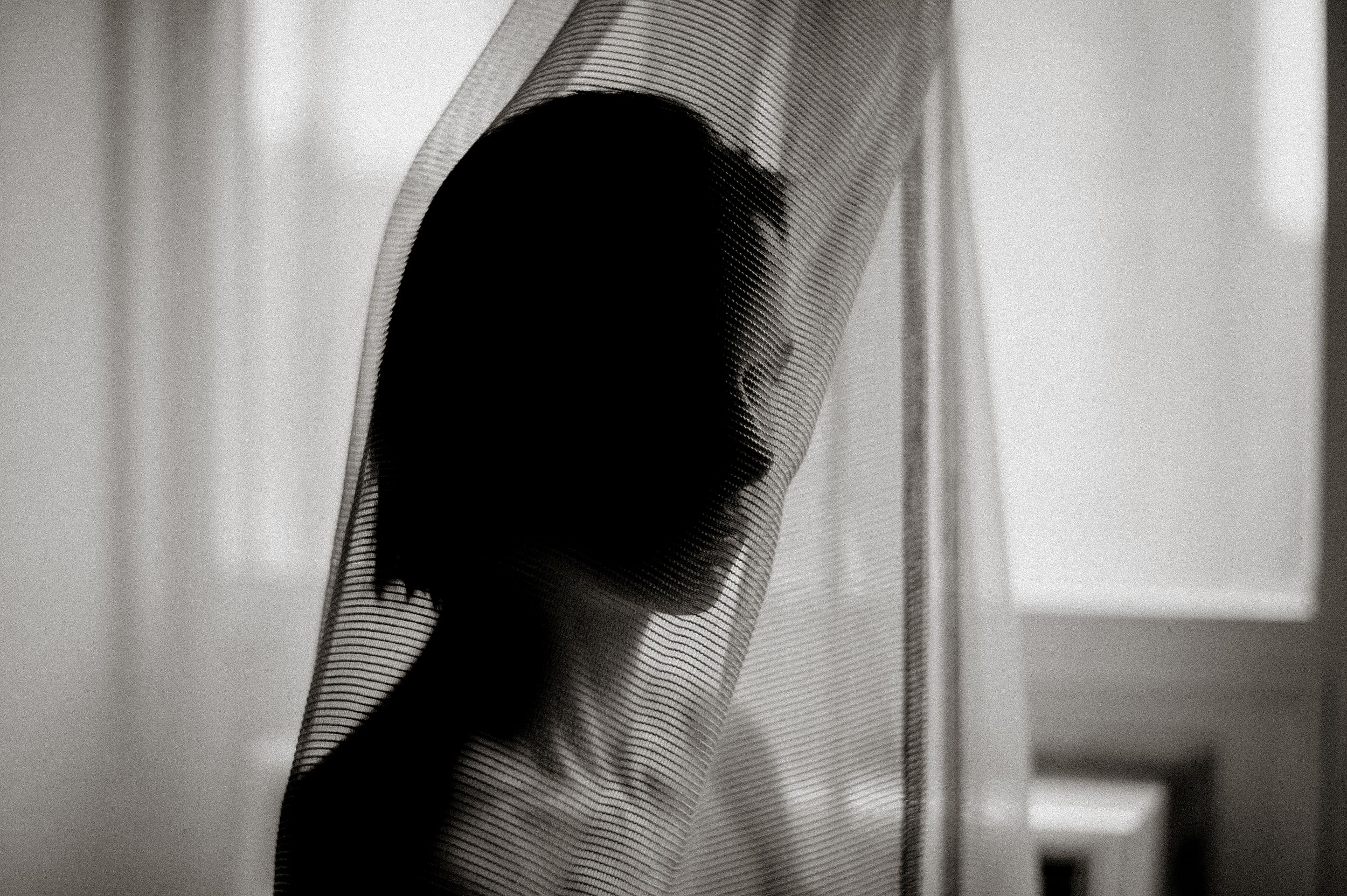 Silhouette of a person with short hair behind sheer curtain in black and white photography.