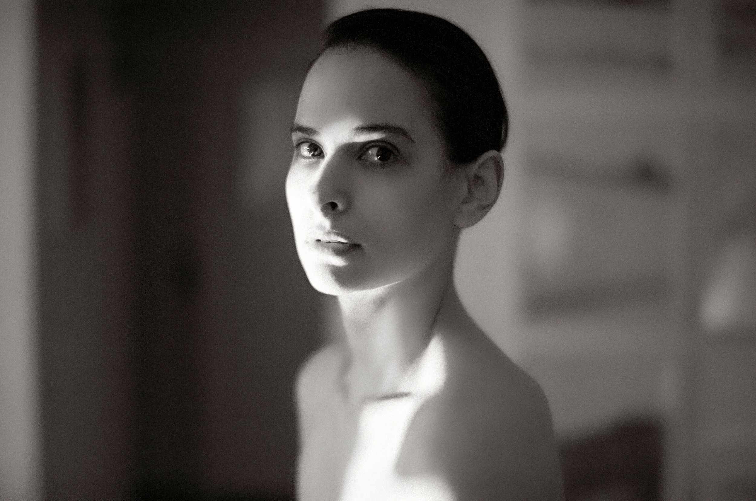 Black and white portrait of a young woman with elegant features and bare shoulders in soft natural light.