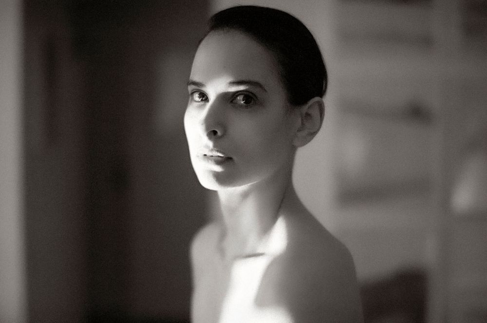 Black and white portrait of a young woman with elegant features and bare shoulders in soft natural light.