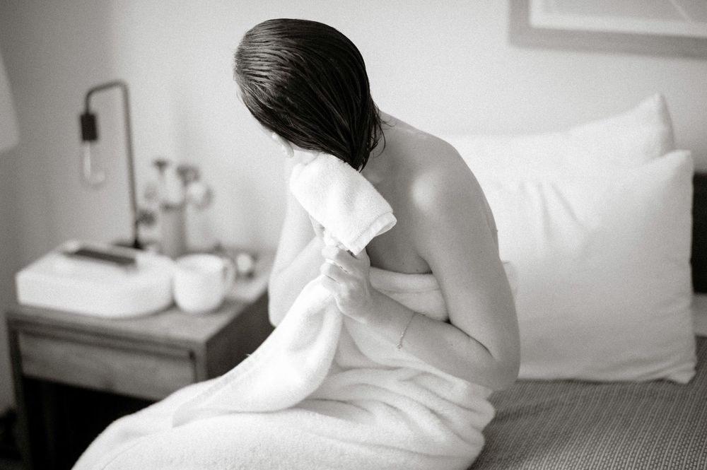 Woman wrapped in white towel sitting on bed drying hair in black and white photo.