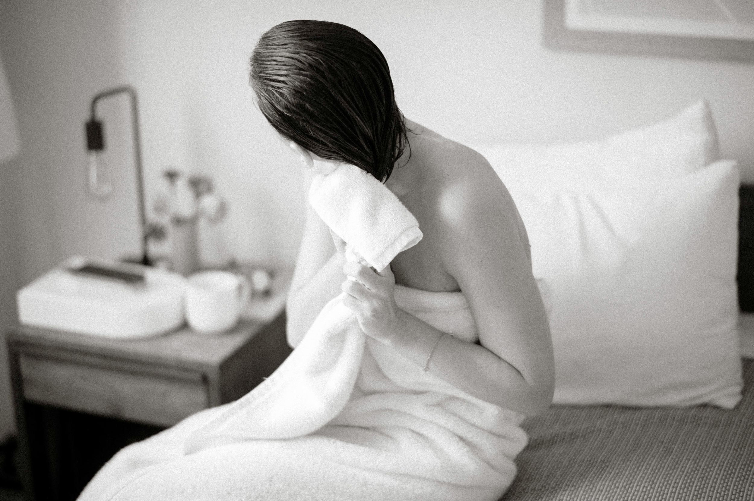 Woman wrapped in white towel sitting on bed drying hair in black and white photo.