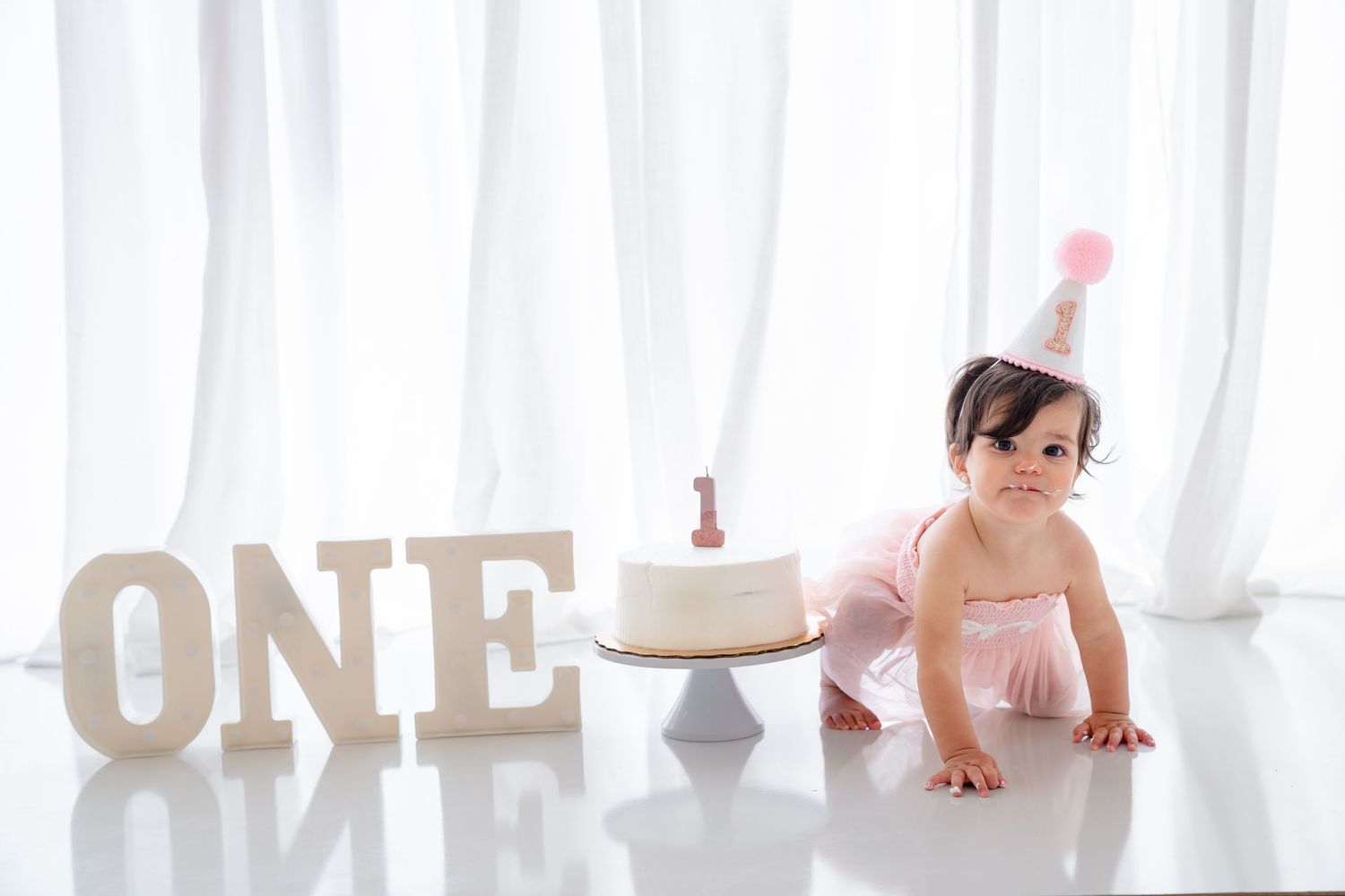 Baby girl in pink dress and party hat crawls near ONE sign and first birthday cake on white floor.