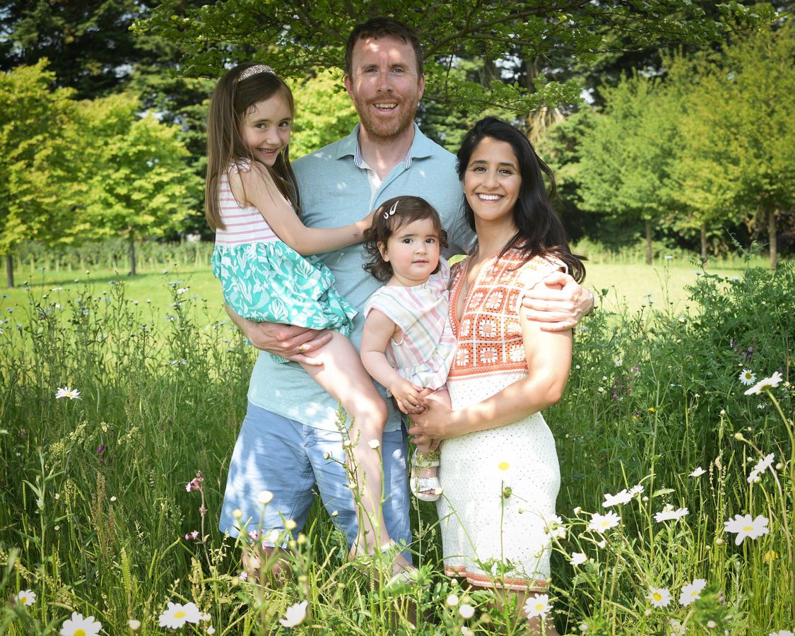 Family of four posing and smiling in a sunny meadow with flowers and trees.