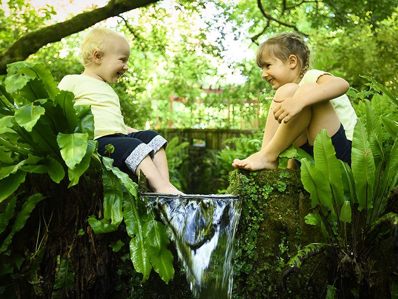 Two toddlers play near a leafy waterfall in a lush green garden.