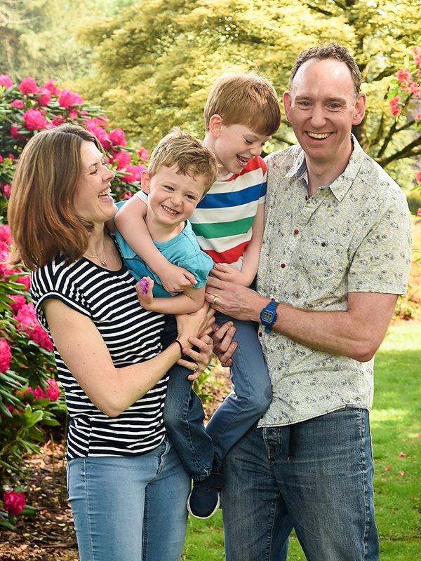 Family smiling outdoors in a garden, man holding child by colourful flowers.