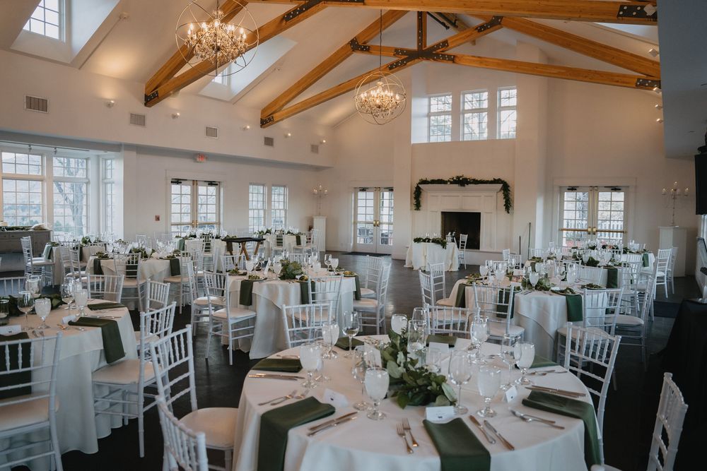 Elegant wedding reception hall with white chiavari chairs, round tables with green runners, and vaulted ceilings with chandeliers.