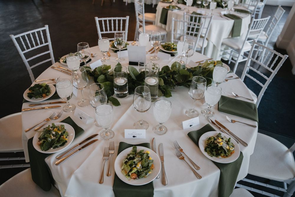 Elegant wedding reception table with white linens, green napkins, gold cutlery, candles, and a lush greenery centerpiece.