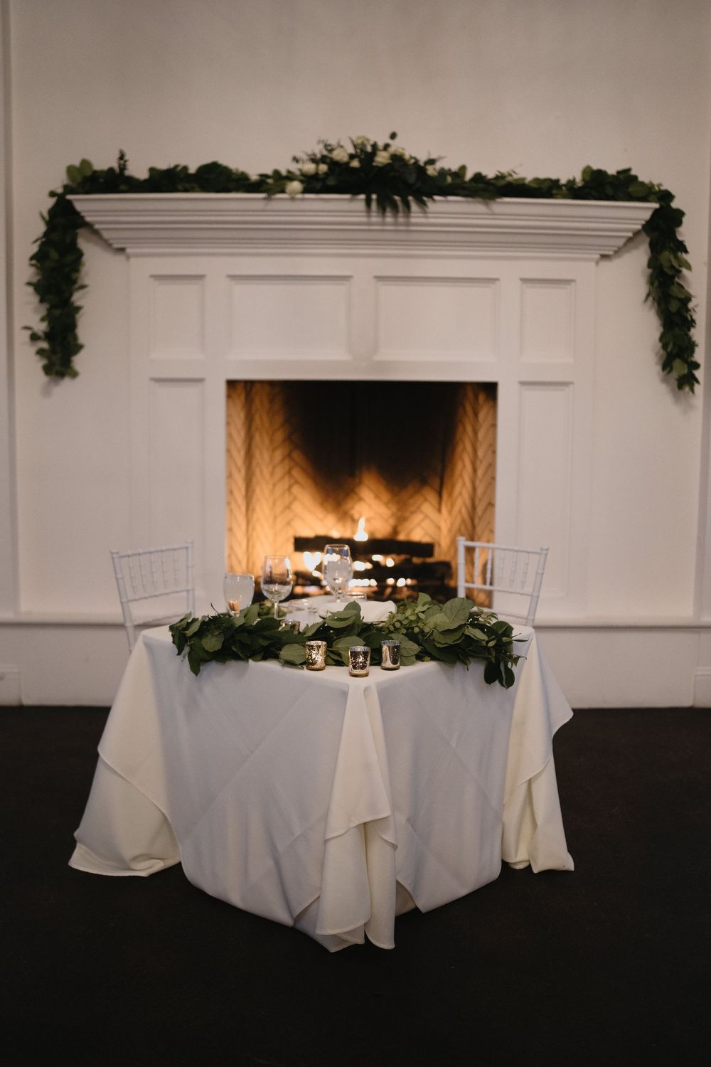 Elegant sweetheart table with white linen and greenery garland set before a glowing fireplace at a wedding reception.