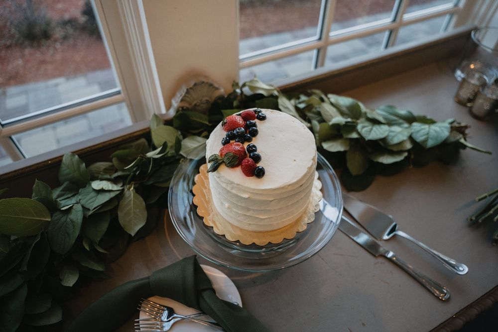 White frosted single-tier wedding cake topped with fresh berries on a glass stand, surrounded by greenery garland.