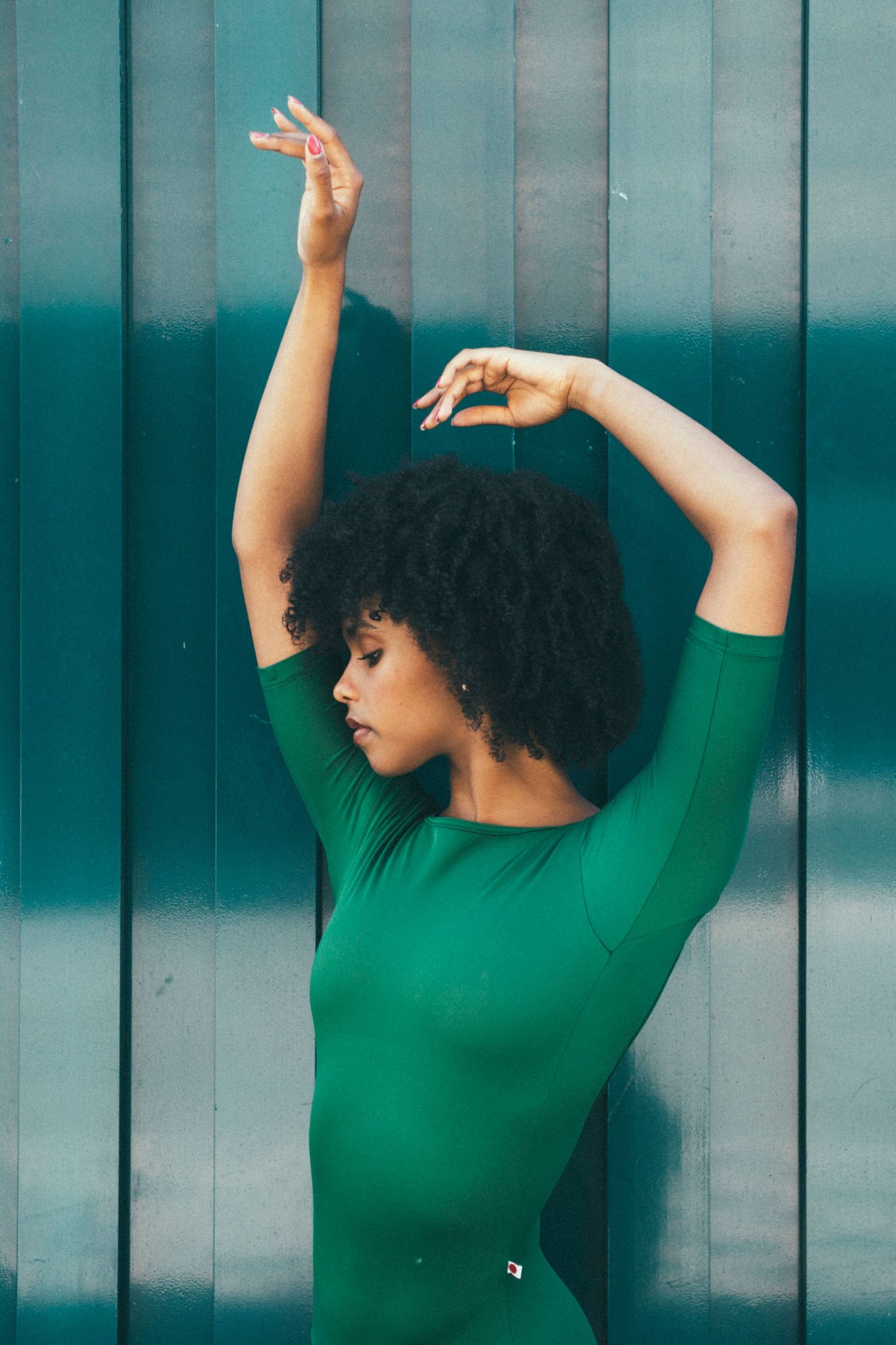 Dancer in green leotard leaning back with arms raised against a teal metal wall with professional Seattle dance photographer