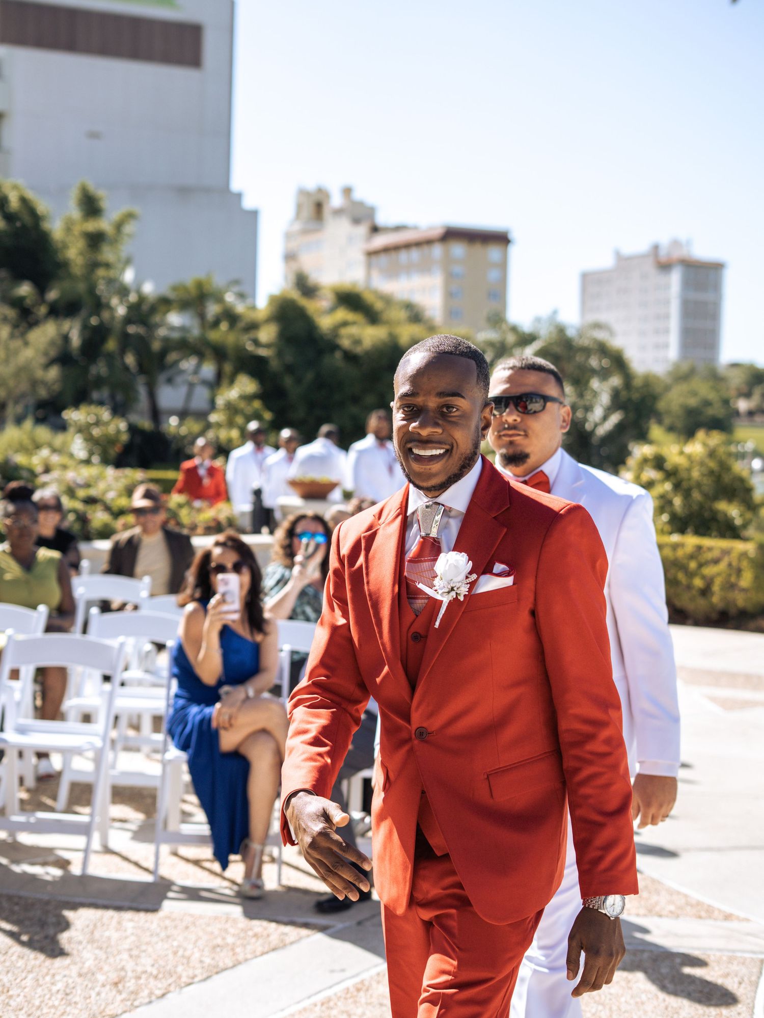 Smiling groom in red suit with boutonniere walks down outdoor wedding aisle with guests seated on white chairs at Hollis Garden in Lakeland