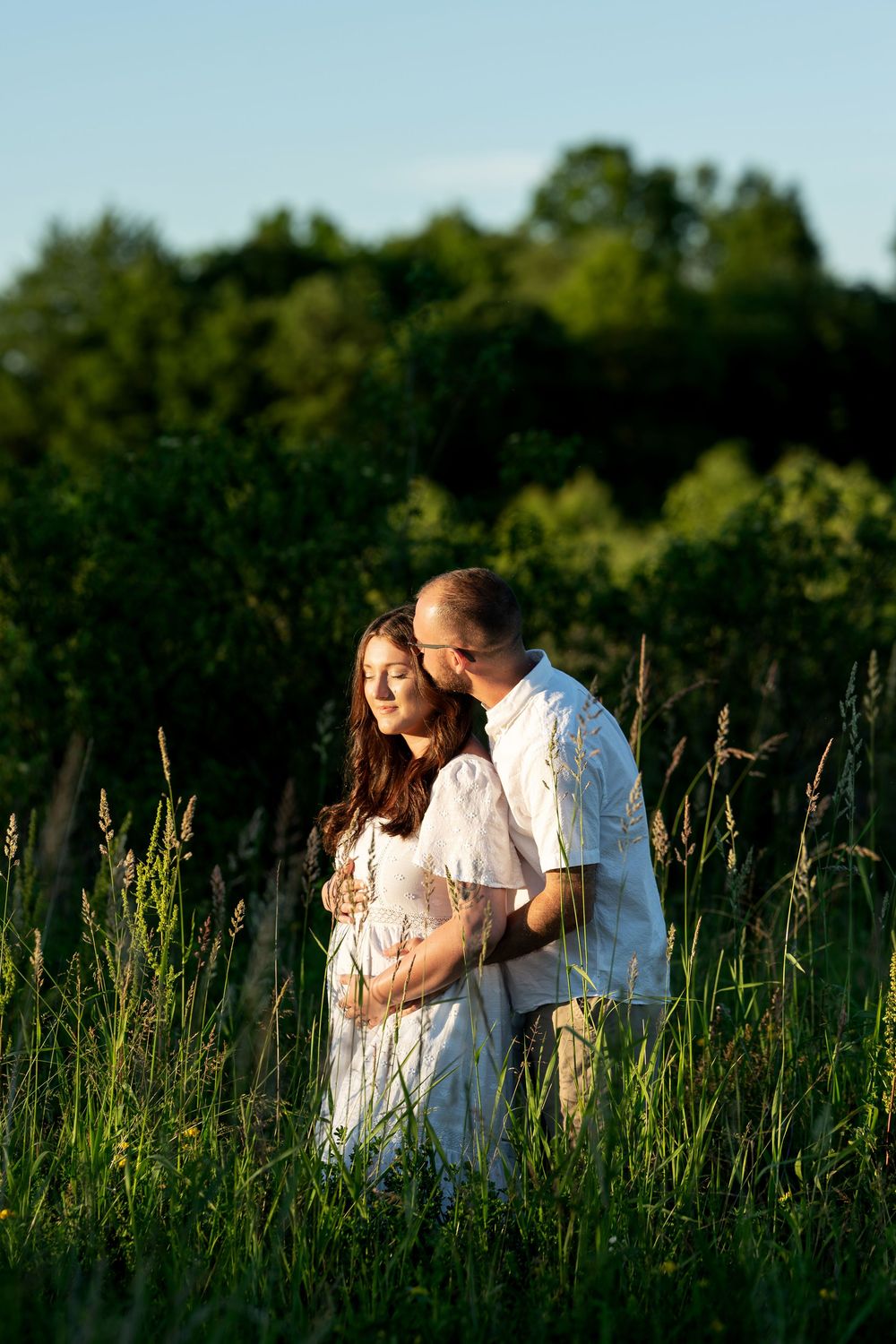 Couple embracing in a sunny green field, pregnant woman in white dress with partner kissing her head.
