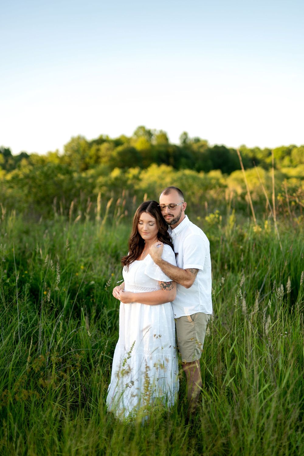 Couple embracing tenderly in tall green grass field, woman in flowing white maternity dress.